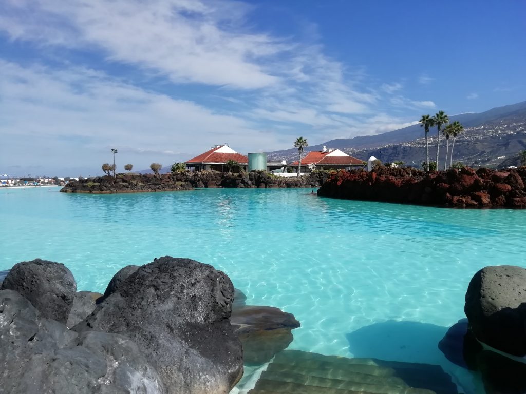 Výhled na modrou vodu akvaparku Lago Martiánez v Puertu de la Cruz na Tenerife. V popředí jsou umělé skály, v pozadí jsou vidět střechy domků a nad nimi se tyčící palmy.